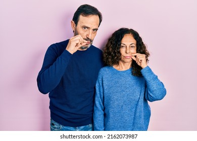 Middle age couple of hispanic woman and man hugging and standing together mouth and lips shut as zip with fingers. secret and silent, taboo talking 