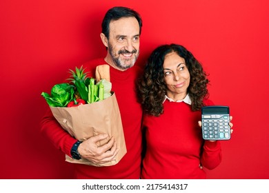 Middle Age Couple Of Hispanic Woman And Man Holding Groceries Bag And Calculator Smiling Looking To The Side And Staring Away Thinking. 