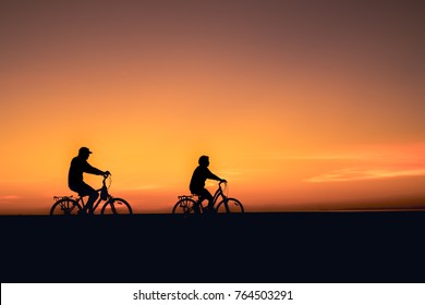 A Middle Age Couple, Riding Their Bicycle At The Beach Bike Path For Better Health.  Bright Beautiful Sunset, Silhouette And Copy Space.