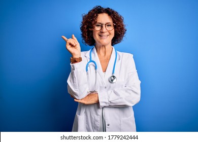 Middle age curly hair doctor woman wearing coat and stethoscope over blue background with a big smile on face, pointing with hand and finger to the side looking at the camera.