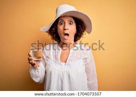 Middle age curly woman on vacation drinking glass of water over isolated yellow background scared in shock with a surprise face, afraid and excited with fear expression