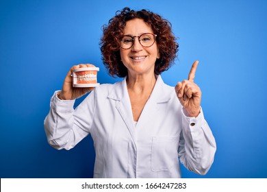 Middle age dentist woman wearing coat holding plastic denture teeth over blue background surprised with an idea or question pointing finger with happy face, number one