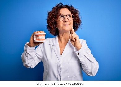 Middle age dentist woman wearing coat holding plastic denture teeth over blue background serious face thinking about question, very confused idea