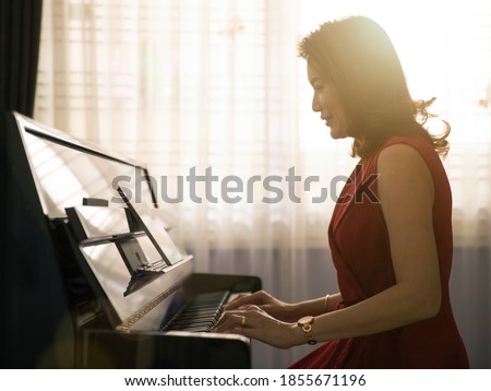 Middle age dignified Asian woman learning and studying new skills how to play the piano via online class from a tablet computer with happiness. Beautiful orange sunlight comes from window in the back. Stock photo © 