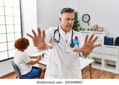 Middle age doctor man at the clinic with a patient afraid and terrified with fear expression stop gesture with hands, shouting in shock. panic concept. 
