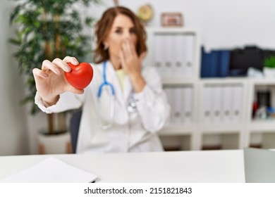 Middle Age Doctor Woman Holding Heart At The Clinic Smiling With Hand Over Ear Listening And Hearing To Rumor Or Gossip. Deafness Concept. 