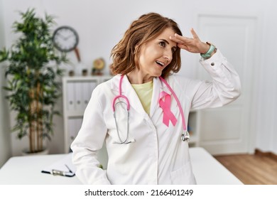 Middle Age Doctor Woman Wearing Pink Cancer Ribbon On Uniform Very Happy And Smiling Looking Far Away With Hand Over Head. Searching Concept. 