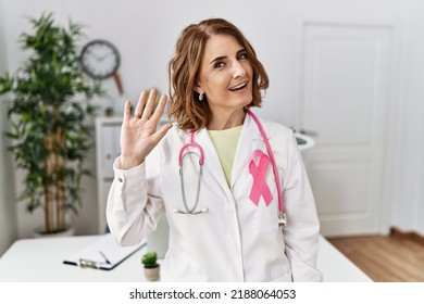 Middle age doctor woman wearing pink cancer ribbon on uniform waiving saying hello happy and smiling, friendly welcome gesture  - Shutterstock ID 2188064053