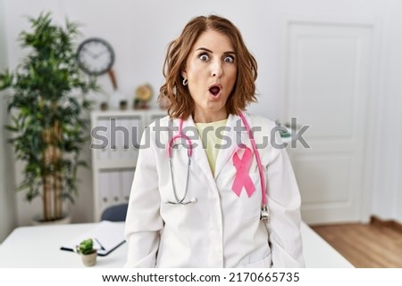Middle age doctor woman wearing pink cancer ribbon on uniform in shock face, looking skeptical and sarcastic, surprised with open mouth 