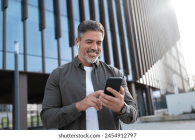 Middle age entrepreneur businessman holding mobile cell phone, watching, listening at office. Latin smiling mature hispanic business man using smartphone cellphone app, earphones for voice call app.  - Powered by Shutterstock