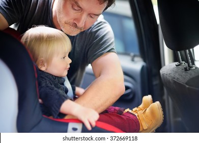 Middle age father helps his toddler son to fasten belt on car seat