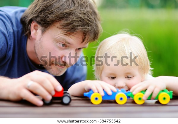 Middle age father with his toddler son\
playing with toy trains outdoors. Fatherhood\
concept