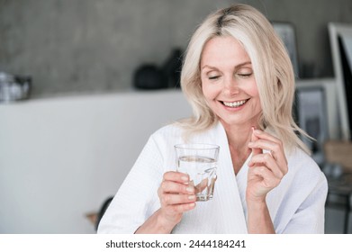 Middle age female holds pill and glass of water in her hands. Woman in menopause taking minerals for skin and health care, boost metabolism. Vitamin A, D, C deficiency prevention treatment. Copy space - Powered by Shutterstock