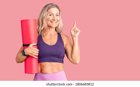 Middle Age Fit Blonde Woman Holding Yoga Mat Surprised With An Idea Or Question Pointing Finger With Happy Face, Number One 