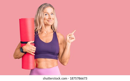 Middle Age Fit Blonde Woman Holding Yoga Mat Smiling Happy Pointing With Hand And Finger To The Side 