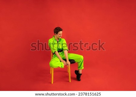 Middle age funny and Cheerful woman wear modern green overalls sitting on chair over red color background. Full length body size photo Female having fun and energetic movement studio shot 