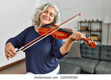 Middle age grey-haired artist woman playing violin standing at home.