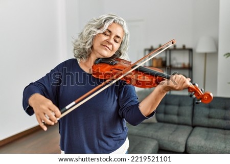 Middle age grey-haired artist woman playing violin standing at home.
