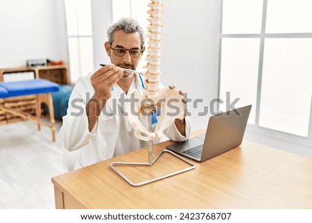 Middle age grey-haired man doctor holding anatomical model of spinal column at rehab clinic