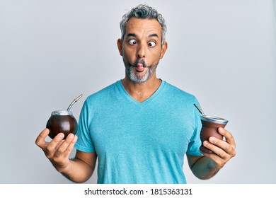 Middle age grey-haired man drinking mate infusion making fish face with mouth and squinting eyes, crazy and comical. 