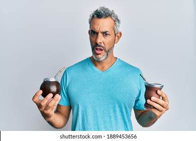 Middle age grey-haired man drinking mate infusion in shock face, looking skeptical and sarcastic, surprised with open mouth 