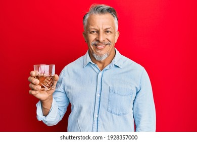 Middle age grey-haired man drinking glass of whisky looking positive and happy standing and smiling with a confident smile showing teeth 
