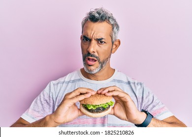 Middle age grey-haired man eating a tasty classic burger in shock face, looking skeptical and sarcastic, surprised with open mouth  - Powered by Shutterstock