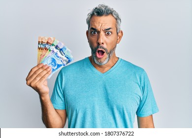 Middle age grey-haired man holding canadian dollars scared and amazed with open mouth for surprise, disbelief face