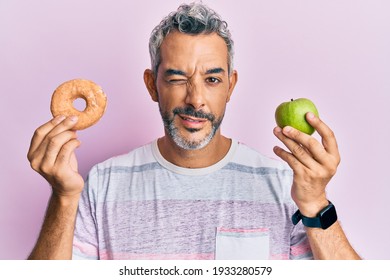 Middle age grey-haired man holding green apple and donut winking looking at the camera with sexy expression, cheerful and happy face. 