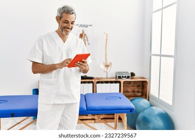Middle age grey-haired man physiotherapist smiling confident using touchpad at rehab clinic