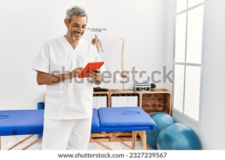Middle age grey-haired man physiotherapist smiling confident using touchpad at rehab clinic