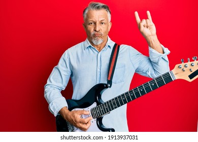 Middle Age Grey-haired Man Playing Electric Guitar Doing Rock Sign Puffing Cheeks With Funny Face. Mouth Inflated With Air, Catching Air. 