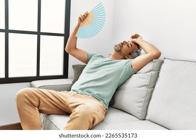 Middle age grey-haired man using handfan sitting on sofa at home