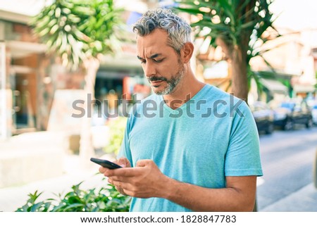 Middle age grey-haired man using smartphone at street of city thinking attitude and sober expression looking self confident 
