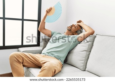 Middle age grey-haired man using handfan sitting on sofa at home
