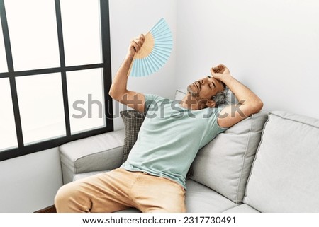 Middle age grey-haired man using handfan sitting on sofa at home