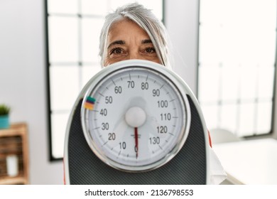 Middle age grey-haired woman covering face with weighing machine at home