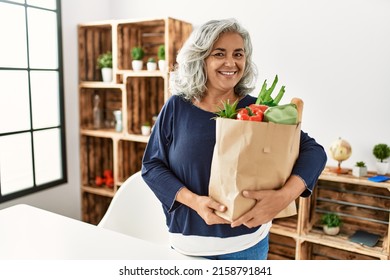 Middle age grey-haired woman holding paper bag with groceries standing at home. - Powered by Shutterstock