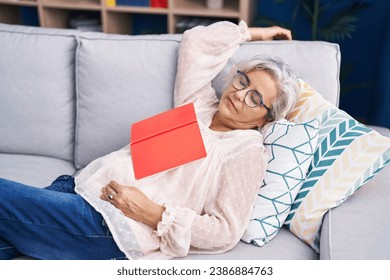 Middle age grey-haired woman holding book lying on sofa sleeping at home - Powered by Shutterstock