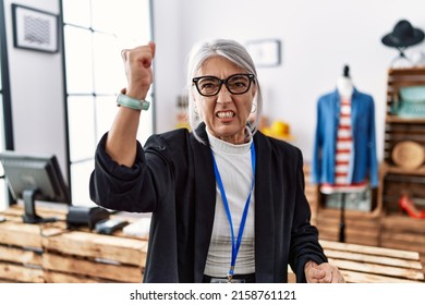 Middle age grey-haired woman working as manager at retail boutique angry and mad raising fist frustrated and furious while shouting with anger. rage and aggressive concept. 