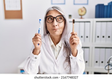 Middle age grey-haired woman working at dentist clinic holding electric and recycled teethbrush smiling looking to the side and staring away thinking. 