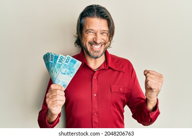 Middle Age Handsome Man Holding 100 Brazilian Real Banknotes Screaming Proud, Celebrating Victory And Success Very Excited With Raised Arm 