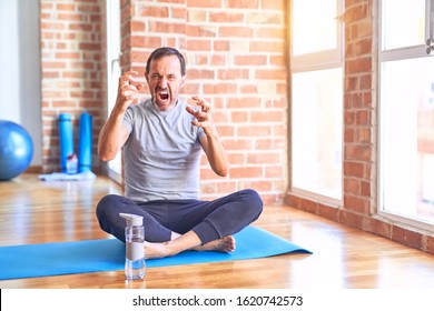 Middle Age Handsome Sportman Sitting On Mat Doing Stretching Yoga Exercise At Gym Shouting Frustrated With Rage, Hands Trying To Strangle, Yelling Mad