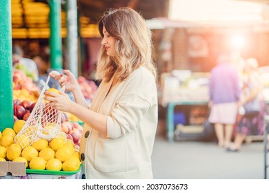 A middle age happy pregnan woman puts a lemon fruits into the mesh bag next to her belly at food market. Photos of fetal growth at 34 weeks pregnancy. Healthy pregnancy diet with vitamin C.