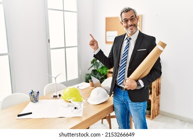 Middle age hispanic business man holding paper blueprints with a big smile on face, pointing with hand finger to the side looking at the camera. 