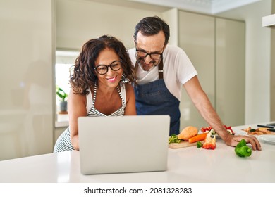 Middle Age Hispanic Couple Cooking And Using Laptop At Kitchen