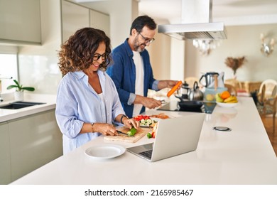 Middle Age Hispanic Couple Cooking And Using Laptop At Kitchen