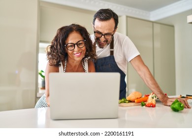 Middle Age Hispanic Couple Cooking And Using Laptop At Kitchen