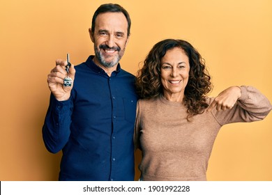 Middle age hispanic couple holding keys of new home pointing finger to one self smiling happy and proud 