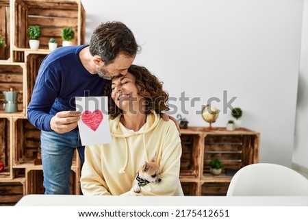 Middle age hispanic couple kissing and holding paper with heart draw at home.